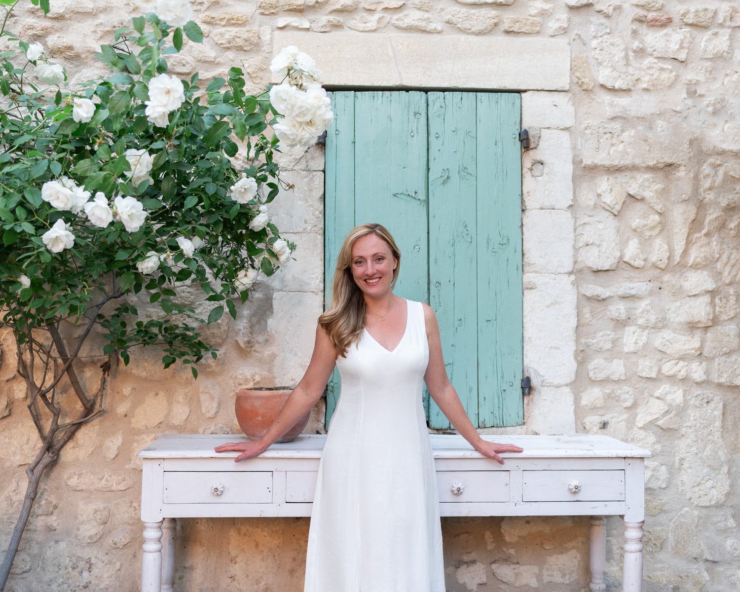 The owner of Countryside Curations, a woman in a white dress standing in front of a stone wall with a turquoise shutter.