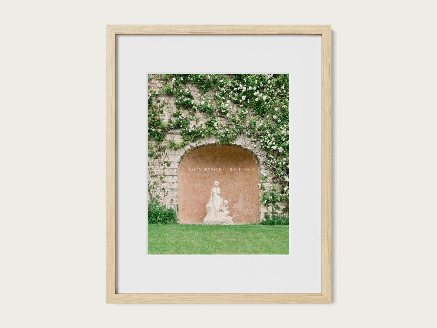 Fine art photograph of a classical garden statue in a stone alcove with climbing greenery and soft white flowers.