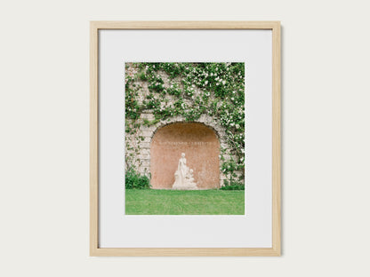 Fine art photograph of a classical garden statue in a stone alcove with climbing greenery and soft white flowers.
