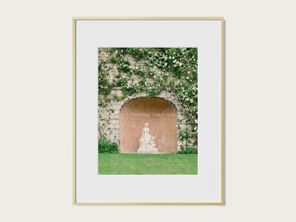 French garden photography print featuring a statue framed by an arched stone wall with lush vines and grass.