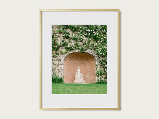 European courtyard photograph of a classical statue within a textured stone alcove covered in climbing plants.